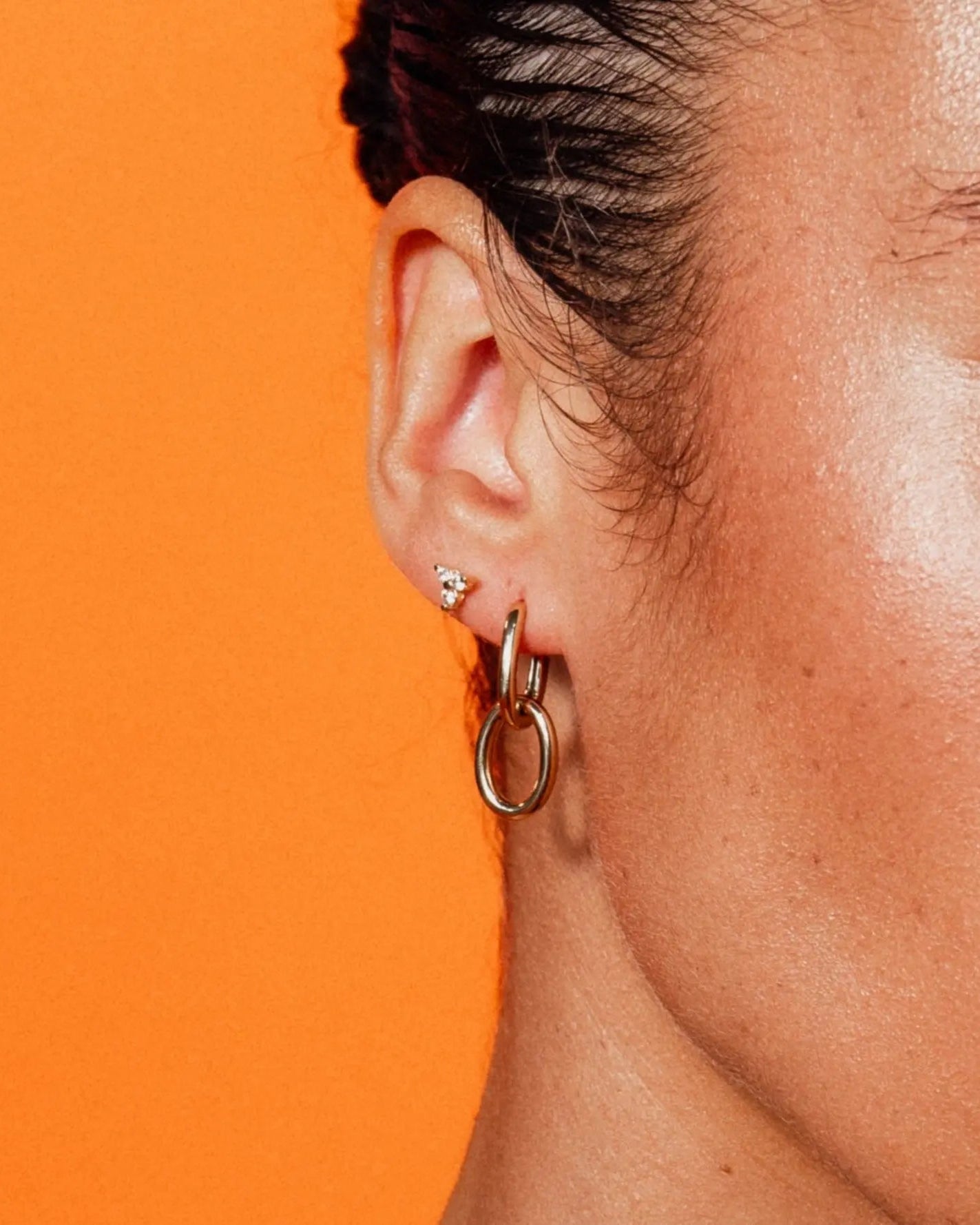 Model wearing Double Paperclip Hoops by IGGI Jewellery – 18k gold-filled interlocking hoops styled on ear with an orange background.