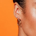 Model wearing Double Paperclip Hoops by IGGI Jewellery – 18k gold-filled interlocking hoops styled on ear with an orange background.