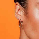 Model wearing Double Paperclip Hoops by IGGI Jewellery – 18k gold-filled interlocking hoops styled on ear with an orange background.