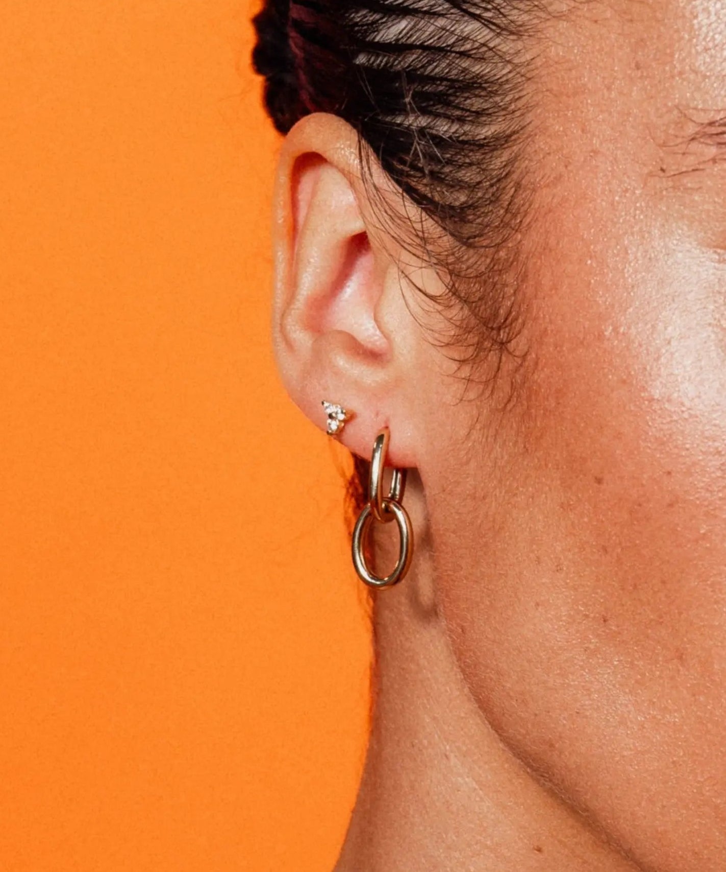 Model wearing Double Paperclip Hoops by IGGI Jewellery – 18k gold-filled interlocking hoops styled on ear with an orange background.